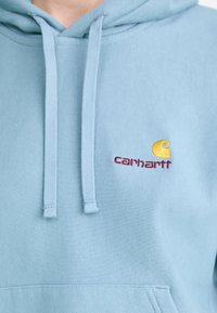Felpa leggera blu chiaro in misto cotone, con tasca frontale, cappuccio con cordino e logo Carhartt ricamato in giallo e bordeaux.
