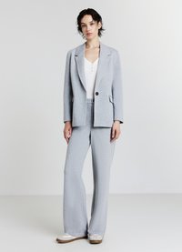Femme se tenant contre un fond uni, portant un blazer gris clair et un pantalon large assorti, avec des baskets blanches et un haut blanc.