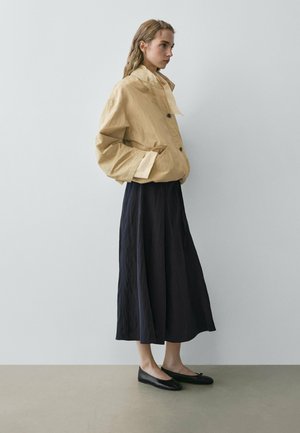 Woman standing sideways wearing a beige oversized jacket, black midi skirt, and black ballet flats against a plain gray background.