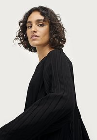 Schwarzes, plissiertes Kleid mit langen Ärmeln und rundem Ausschnitt, gefertigt aus weichem Stoff und mit einer lockeren Silhouette. Kopf des Models leicht gedreht.