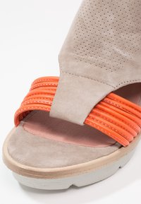 Sandale beige avec partie supérieure perforée et trois brides orange vif cousues sur les orteils sur une semelle blanche épaisse.