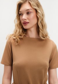 Blondynka z falującymi włosami patrzy w lewo, ubrana w brązowy t-shirt z krótkim rękawem na jednolitym białym tle.