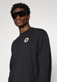 Sudadera negra con un parche de logo redondo en el pecho, cuello redondo y mangas largas; llevada con grandes gafas de sol negras.