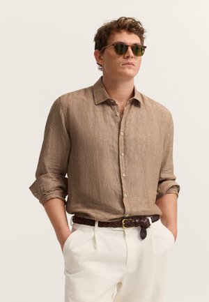 Mann trägt beige Leinenhemd mit hochgekrempelten Ärmeln, weiße Hose, geflochtener brauner Gürtel und Sonnenbrille, Hände lässig in den Taschen vor beigem Hintergrund.