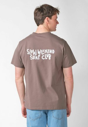 Mees, kes on seljaga poole keeratud, kannab pruunikat T-särki, millel on tagaküljel kiri "SLOW WEEKEND SURF CLUB", ning helesiniseid teksapükse.