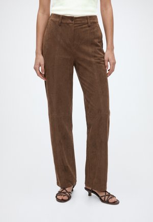 Mujer que lleva pantalones marrones de ante rectos y de talle alto con costura frontal, combinados con camiseta blanca y sandalias negras de tiras con tacón bajo.