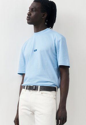 Άνδρας με σκούρο δέρμα και dreadlocks που φοράει ανοιχτό μπλε μπλουζάκι Hugo, κρυμμένο μέσα σε άσπρο παντελόνι με μαύρη ζώνη, κοιτάζει αριστερά σε απλό φόντο.