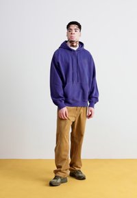 Sudadera morada, pantalones de pana caqui y zapatillas de deporte verde oscuro. La sudadera tiene un bolsillo frontal y un pequeño logo; los pantalones son ajustados.