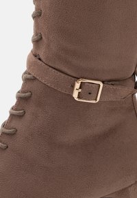 Hnědé boty z umělé semišové kůže s detaily šněrování a zlatým akcentem sponky na kotníku pro bezpečné zapnutí.