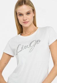 Hvid t-shirt med korte ærmer, der har et sølv rhinesten print med ordet "Lingo" hen over brystet, samt en glat bomuldsstruktur.