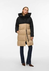 Pufjakas ar melnu augšdaļu un bēšu apakšdaļu, ar kapuci, rāvējslēdzēju un sānu kabatām, kas izgatavots ar quilted tekstūru.