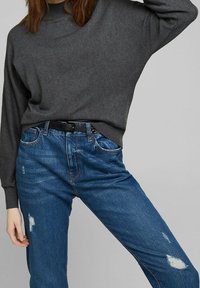 Grijze, langwerpige trui met lange mouwen, gecombineerd met blauwe jeans met versleten plekken en een zwarte riem. De trui heeft een ronde halslijn en een ontspannen pasvorm.