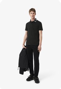 Svart polotröja med kontrasterande vit text på kragen, tillsammans med svarta byxor och svarta sneakers. Modellen håller en svart jacka.