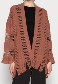 Otwarto-splotowy poncho w kolorze rdzawym, z dekoltem w kształcie V, bogatymi wzorami, frędzlami na dolnym brzegu i luźnymi rękawami. Całość wykonana z teksturowanego materiału.