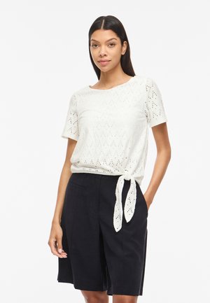 Žena oblečená v bielom krátkoročnom topu s dierkami a bočným viazaním a čiernych šortkách po kolená, stojaca pred jednofarebným bielym pozadím.