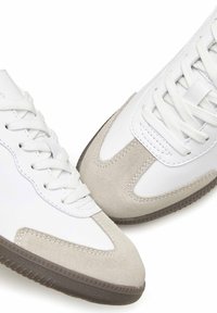 Weiße Sneakers mit einer Mischung aus glattem Leder und beigen Wildlederakzenten, flachen weißen Schnürsenkeln und einer strukturierten grauen Gummisohle.