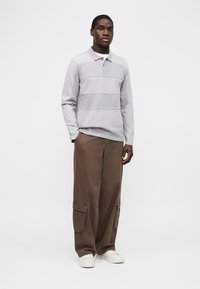 Homme debout portant un polo à manches longues gris clair, un pantalon cargo marron à jambes larges et des baskets blanches sur un fond uni.