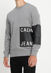 Šedá mikina s černou ekokůží nášivkou, na které je vytištěn nápis "CALVIN JEANS" v tučných bílých písmenech. Rázy na rukávech a kulatý výstřih.