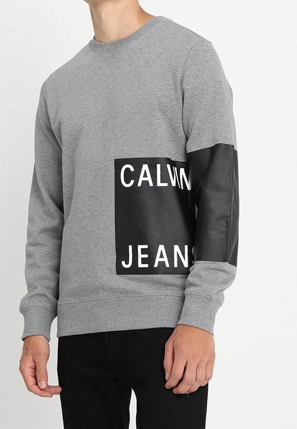Šedá mikina s černou ekokůží nášivkou, na které je vytištěn nápis "CALVIN JEANS" v tučných bílých písmenech. Rázy na rukávech a kulatý výstřih.
