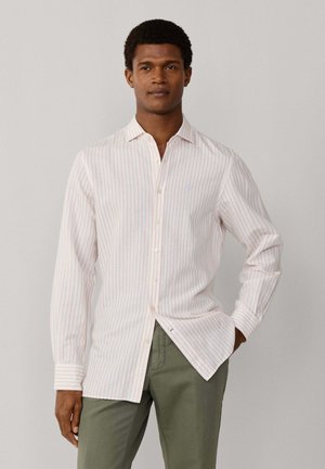 Homme portant une chemise blanche à manches longues avec de fines rayures verticales rouges et un pantalon vert olive, debout avec une main dans la poche.