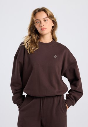 Jonge vrouw met lichtbruin haar in een donkerbruine sweatshirt en bijpassende broek, handen in de zakken, neutrale achtergrond.