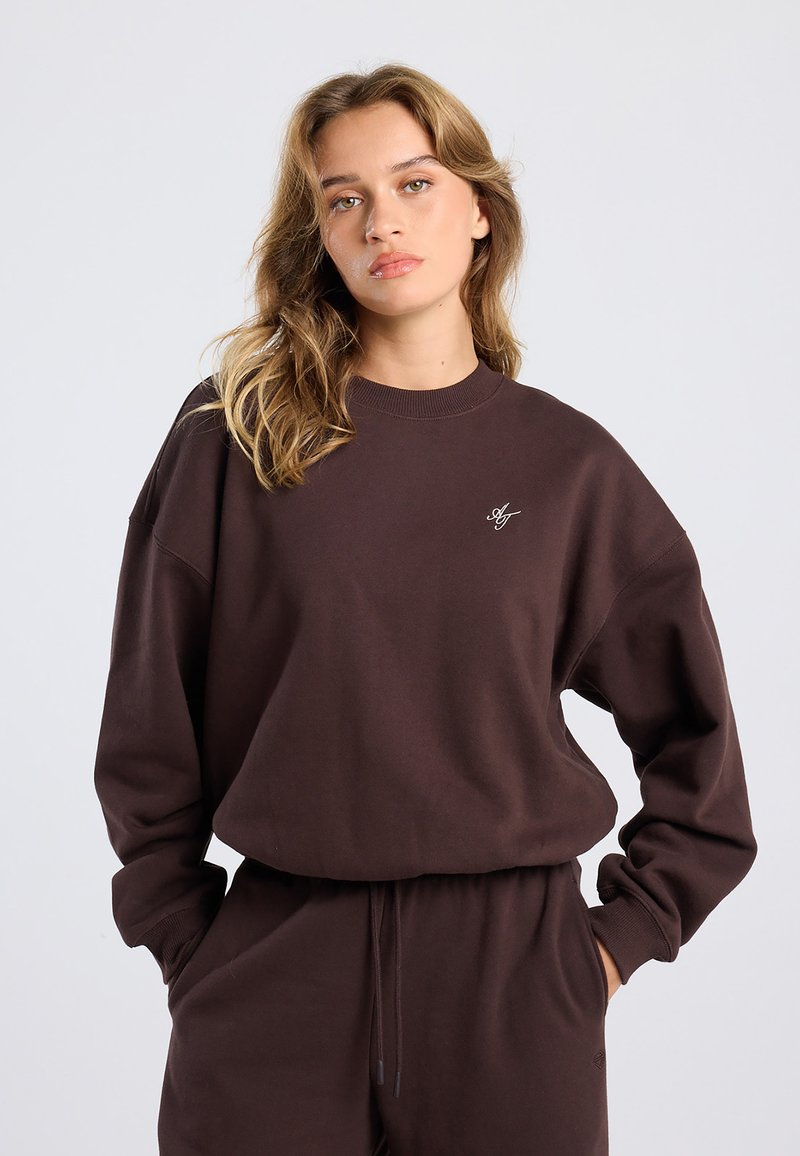 Young woman with light brown hair wearing a dark brown sweatshirt and matching pants, hands in pockets, neutral background.