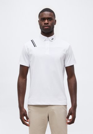 Hombre de pie mirando hacia adelante con una camiseta polo blanca con dos rayas negras en el hombro derecho y pantalones beige.