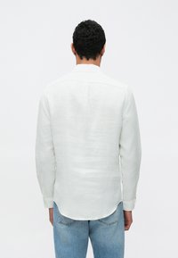 Camisa branca de linho de manga comprida com corte reto, punhos abotoados e um ligeiro brilho. Combinada com jeans azul claro, mostrando um ajuste descontraído.