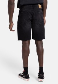 Shorts en denim noir avec un ourlet effiloché. Comprend deux poches arrière et une étiquette en cuir beige à la taille. Portés avec des chaussettes noires et des chaussures.