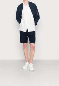 Marineblaue Bomberjacke, weißes Leinenhemd mit Knöpfen, dunkle Shorts und weiße Turnschuhe. Schlichtes Design mit Reißverschlusstasche an der Jacke.