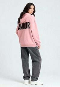Felpa rosa con "AMOUR" in bold lettere nere sulla schiena, abbinata a jeans grigi larghi e sneakers bianche. Tessuto morbido e vestibilità comoda.