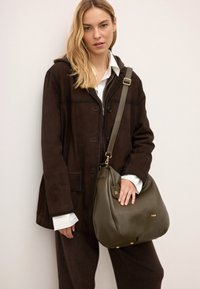 Barna velúr kabát, fehér galléros ing, zöld bőr kézi táska arany kiegészítőkkel, sima textúra, laza forma, keresztben hordva.