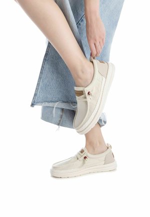 Zapatos sin cordones de color beige, hechos de lona con un acabado texturizado. Presentan una suela trenzada y un pequeño lazo en el talón, con acentos mínimos.