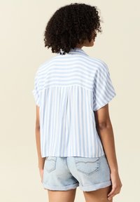 Kortärmad skjorta i ljusblå och vita ränder, med en gathered backdetalj, framtagen i ett lätt tyg. I kombination med denimshorts.