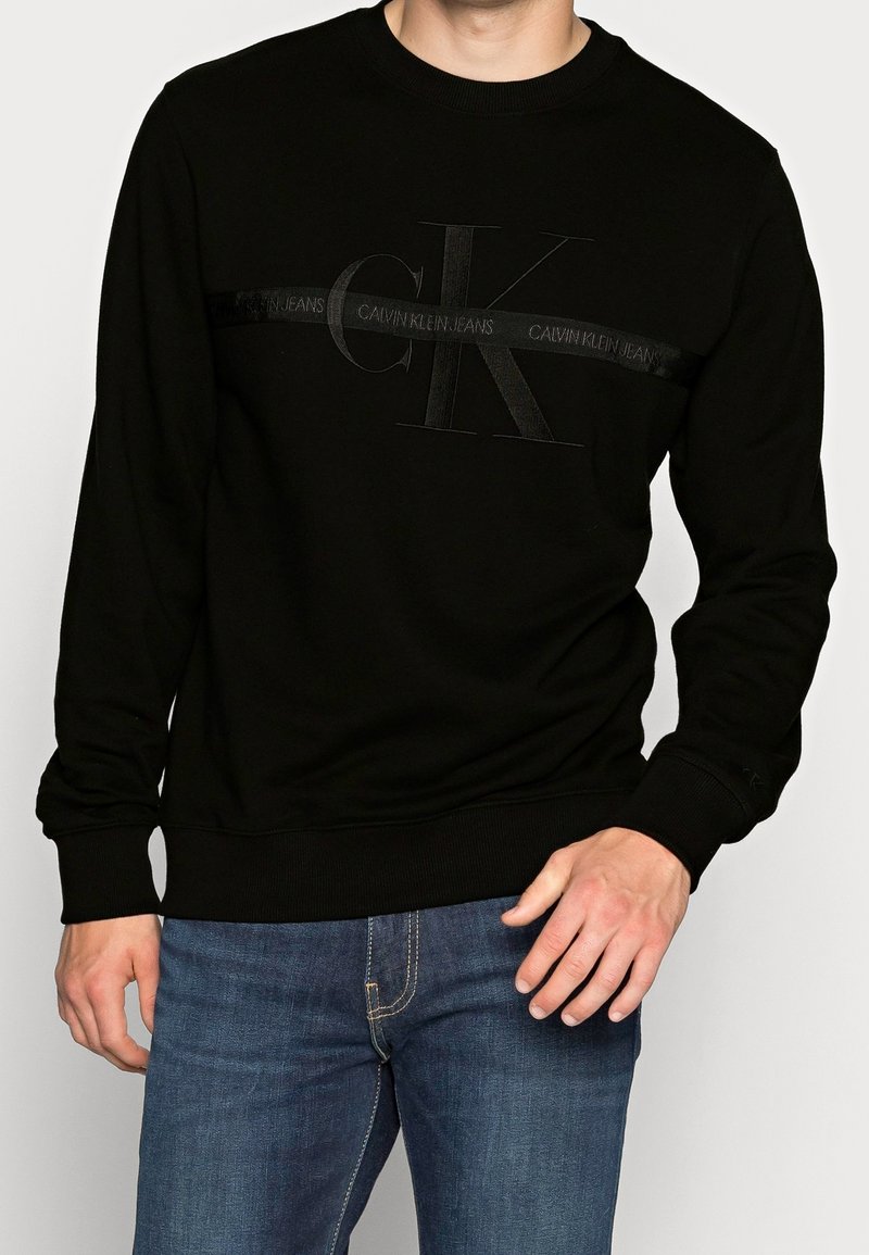 Homme portant un sweat-shirt noir Calvin Klein avec logo CK embossé et texte horizontal, associé à un jean bleu.