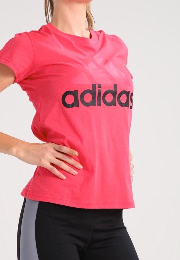 Femme portant un t-shirt Adidas rose vif avec un logo noir, les mains sur les hanches, associé à des leggings noirs et gris sur un fond uni.