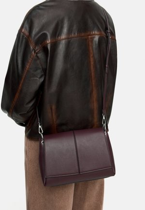 Osoba ubrana w ciemnobrązową skórzaną kurtkę i beżowe spodnie, niosąca usztywnioną, ciemnofioletową torbę na ramię z srebrnymi zdobieniami.