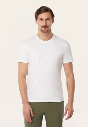 Mann mit kurzen braunen Haaren und Schnurrbart, trägt ein schlichtes weißes T-Shirt und olivgrüne Hose, steht vor einem hellen Hintergrund.