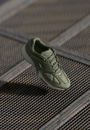 Green athletic shoe with textured fabric upper, lace closure, and white sole. Features curved design lines and subtle branding details.