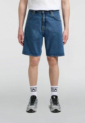 Persona con pantaloncini di jeans blu, calzini bianchi con scritta nera, scarpe da ginnastica argentate e nere, in piedi su un pavimento grigio.