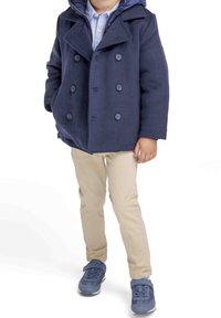 Marinblå sjömansrock med dubbelknäppta knappar, bärs över en ljusblå skjorta och kombineras med beige byxor och blå sneakers.