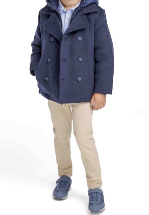 Marineblå peacoat med dobbeltknaplukning, båret over en lyseblå skjorte og parret med beige bukser og blå sneakers.