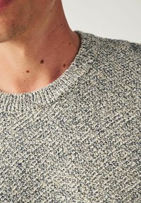 Gros plan sur un cou et une épaule portant un pull en tricot texturé aux tons beige et bleu foncé.