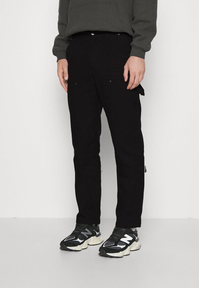 Pantalon cargo noir avec poches latérales, coupe droite et garnitures en ton cuivre, associé à des baskets athlétiques noires et blanches.
