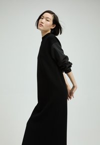 Schwarzes Kleid mit langen, gepufften Ärmeln, aus einem strukturierten Stoff gefertigt. Lockere Passform, mittellanger Stil, der ein minimalistisches Design ohne Muster aufweist.