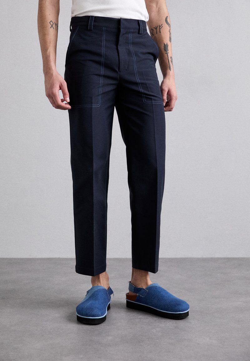 Navy blue tailored pants with contrasting light blue stitching, straight cut, and a flat front. Paired with blue slip-on shoes featuring a brown heel.