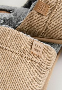 Pantofole intrecciate di colore beige con un aspetto testurizzato, bordo beige e fodera in pelliccia grigia. Etichetta in pelle marrone sulla parte superiore.