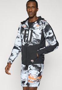 Μαύρο και λευκό σχέδιο στο μπουφάν με φερμουάρ μπροστά και κουκούλα, συνδυασμένο με σορτς που ταιριάζουν και έχουν πορτοκαλί λεπτομέρειες και γραφικά στοιχεία.