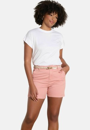 Moteris su garbanotais plaukais, vilkinti baltą trumpomis rankovėmis palaidinę ir rožinius šortus su austu diržu, stovinti viena ranka kišenėje.