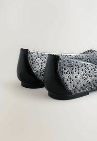 Mustat ja hopeiset tasapohjaiset kengät, joissa on teksturoitu päällinen, jossa on erikokoisia kristalleja, pyöreä kärki ja matala musta korko. Silkkiset nahkaaksentit.